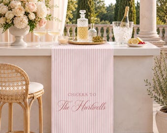 May include: A light pink and white vertical striped table runner featuring the text "CHEERS TO The Hartwells" in a dark pink script font. The runner is draped over a white counter with a glass decanter, a bowl of olives, and a champagne bucket.