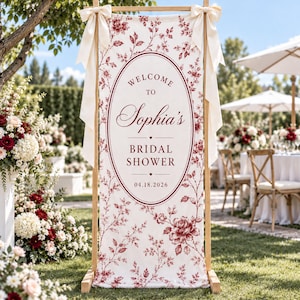 May include: A vertical rectangular fabric banner featuring a red floral pattern on a white background, suspended from a wooden frame with cream ribbons. The centre displays an oval frame with the text Welcome to Sophia's Bridal Shower 04.18.2026.