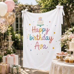 May include: A white fabric birthday banner featuring a colorful illustration of a cake with three candles and the text Happy birthday Ava in a rainbow gradient font, accented with small stars and dots, suspended from a black metal frame.