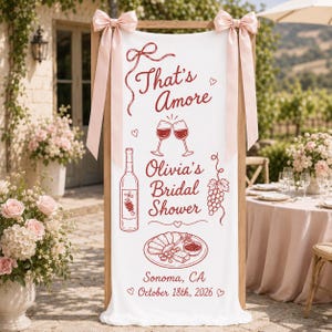 May include: A white vertical fabric banner featuring red line art of a wine bottle, two clinking glasses, a grape cluster, and a charcuterie board. The text reads That's Amore, Olivia's Bridal Shower, Sonoma, CA, and October 18th, 2026. Two light pink bows are attached to the top corners.