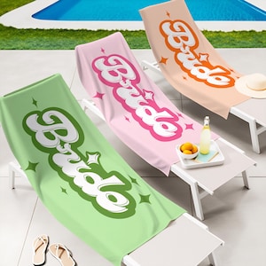 May include: Three beach towels in green, pink, and peach, each with the word "Babe" in a retro font. The towels are on white lounge chairs near a pool. A small table holds a tray with a bottle, lemons, and a hat.