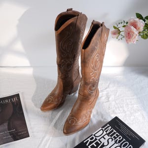Puede incluir: Un par de botas de vaquero marrones con costuras blancas decorativas. Las botas tienen puntas puntiagudas y un tacón de altura media. Las botas se exhiben sobre una superficie blanca con una revista y flores en el fondo.