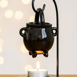 May include: A black ceramic cauldron-shaped oil warmer with a handle and two side handles, suspended from a black metal stand. A lit tea light candle sits below the cauldron, casting a warm glow. The product is designed for aromatherapy.