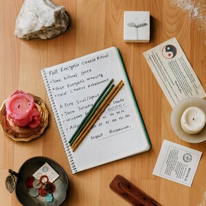 May include: An overhead shot of a spiritual wellness setup on a wooden surface. A notebook with handwritten text, candles, incense, and decorative items are arranged. The notebook reads "Full Energetic Cleanse Ritual". A pink candle sits in a gold dish.