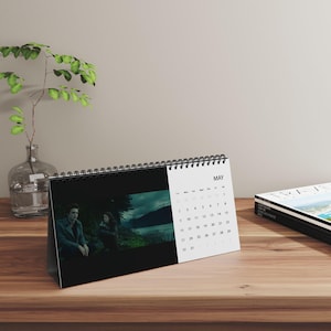 Op de afbeelding: Een bureaukalender met een zwarte spiraalbinding die de maand mei weergeeft. De linkerkant toont een filmscène met twee figuren in een donker buitenlandschap, terwijl de rechterkant een standaard kalenderraster voor de maand mei laat zien.
