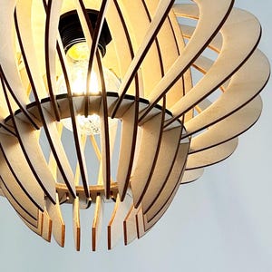 Może przedstawiać: Drewniana lampa wisząca o unikalnym, warstwowym designie. Lampa wykonana jest z jasnego drewna i posiada szereg zakrzywionych, zazębiających się elementów, które tworzą efekt rzeźbiarski. Żarówka jest widoczna pośrodku, emitując ciepłą poświatę.