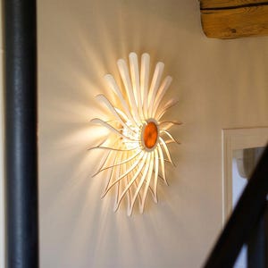 May include: A wall-mounted light fixture featuring a sunburst design with curved, white wooden slats radiating from a central circular orange core. The lamp emits a warm, glowing light that casts soft shadows against the surrounding wall surface.