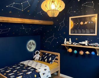 May include: A bedroom featuring dark blue walls with gold constellation decals, a wooden bed frame with rocket-patterned bedding, a moon wall decal, a wooden pendant light, a solar system poster, toy spaceships, and a planet-shaped area rug.