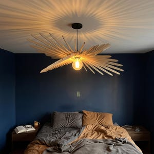 Peut inclure: Un luminaire suspendu au plafond doté d'une ampoule centrale entourée de nombreuses fines lattes de bois clair disposées en un motif radial en forme d'ailes, projetant des ombres distinctes et allongées sur la surface du plafond.