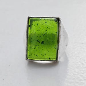 May include: A silver ring featuring a rectangular, lime green stone set in the center. The stone has a smooth, polished surface with small, dark, irregular flecks distributed throughout its translucent, vibrant green body.