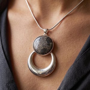 May include: A silver necklace featuring a round pendant with a dark, speckled stone set above a smooth, polished silver crescent shape. The pendant hangs from a thin, flexible silver snake chain that rests against the collarbone area.