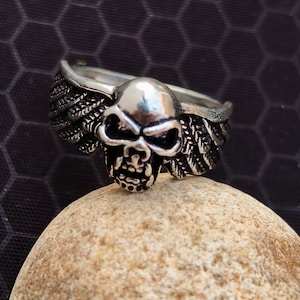 May include: A silver skull ring with detailed wings. The skull has eye sockets and a nose. The ring is displayed on a light brown stone, with a dark background. The ring is likely made of metal.