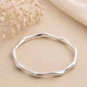 May include: A polished silver bangle bracelet featuring a continuous, smooth, undulating wave pattern. The circular metal band has a reflective, metallic finish and a consistent thickness throughout its entire circumference.