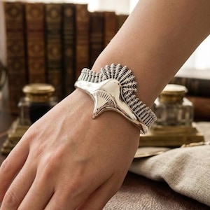 May include: A silver cuff bracelet with a detailed, textured design. The bracelet features a central stylized element and woven-like detailing. The cuff is shown on an arm, showcasing the jewelry's design and craftsmanship. The bracelet is a piece of wearable art.