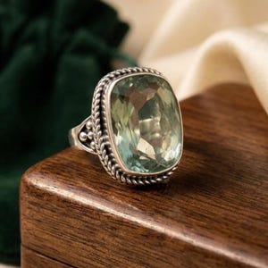 May include: A silver ring featuring a large, rectangular, faceted light green gemstone. The setting is framed by a decorative twisted rope border and small silver bead accents along the sides of the band, showcasing a polished metallic finish.