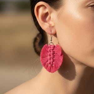 Puede incluir: Primer plano de un pendiente de macramé rosa. El pendiente presenta un diseño central tejido con una franja de hilos rosas. Cuelga de un gancho plateado. El fondo está borroso, enfocando el pendiente.
