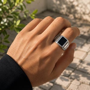 May include: A silver-toned ring featuring a rectangular black stone inlay set within a metallic band. The band displays a textured, grooved pattern along the sides of the setting, providing a distinct contrast to the smooth, dark central stone.