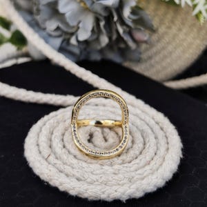 Puede incluir: Un anillo dorado con un diseño único. El anillo presenta una banda horizontal central y una forma ovalada en la parte superior, ambas incrustadas con pequeñas piedras preciosas transparentes. El anillo se muestra sobre una cuerda blanca enrollada sobre un fondo oscuro.