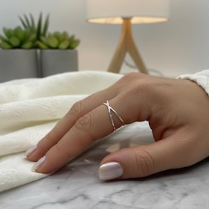 Pode incluir: Um anel prateado com um design cruzado é usado em um dedo. O anel tem um estilo simples e elegante. O fundo inclui tecido de renda branco, uma pequena planta em vaso e uma lâmpada.