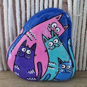 Pode incluir: Uma pedra lisa e arredondada pintada com um fundo azul vibrante apresentando quatro gatos de desenho animado. Os gatos são coloridos em tons de rosa, roxo, azul-petróleo e azul claro, cada um com olhos circulares, bigodes e listras.
