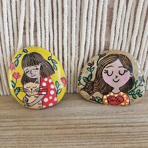 Puede incluir: Dos piedras pintadas a mano con ilustraciones de personas. La piedra izquierda tiene un fondo amarillo con una persona con camisa roja de lunares abrazando a un gato. La piedra derecha es beige con una persona con camisa naranja de lunares.