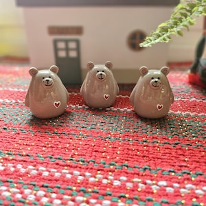Könnte beinhalten: Drei Keramik-Bärenfiguren mit glänzendem Finish. Jeder Bär ist hellbraun mit einem lächelnden Gesicht, kleinen Ohren und einem roten Herz auf der Brust. Sie sind auf einer rot-grün gemusterten Oberfläche angeordnet.