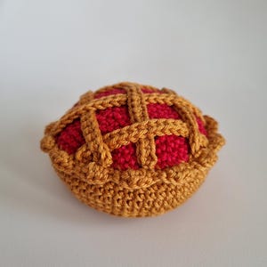 Peut inclure: Une tarte au crochet avec une croûte en treillis. La garniture de la tarte est rouge et la croûte est de couleur brun doré. La tarte est ronde et a un bord décoratif.
