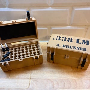 Könnte beinhalten: Zwei rechteckige Aufbewahrungsboxen in Sandfarbe mit schwarzen Verschlüssen. Eine Box ist geöffnet und zeigt ein Raster aus runden Fächern sowie einige kleine Teile mit weißen Spitzen. Der Deckel der geschlossenen Box trägt die Aufschrift .338 LM und A. BRUNNER.