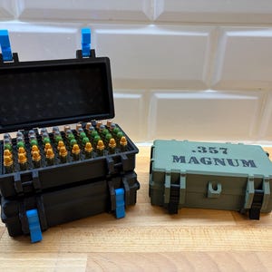 Könnte beinhalten: Zwei schwarze Aufbewahrungsboxen mit blauen Verschlüssen, eine davon offen und mit Munition gefüllt. Eine zweite, geschlossene, grüne Box ist mit ".357 MAGNUM" beschriftet. Die Boxen stehen auf einer Holzoberfläche.