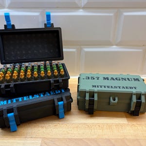 Könnte beinhalten: Drei schwarze und grüne Aufbewahrungsboxen mit blauen Akzenten. Eine Box ist geöffnet und zeigt Munition. Auf den Boxen steht der Text ".357 MAGNUM" und "MITTELSTAEDT". Die Boxen stehen auf einer Holzoberfläche.