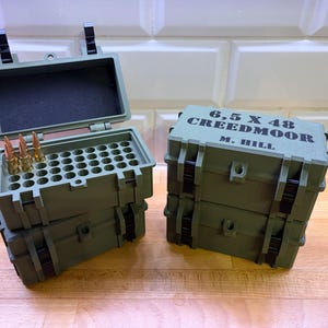 Könnte beinhalten: Zwei olivgrüne Munitionsaufbewahrungsboxen. Eine Box ist geöffnet und zeigt ein Raster von Löchern und Kugeln. Die andere Box ist geschlossen, mit dem Text "6,5 X 48 CREEDMOOR M. HILL" auf dem Deckel. Beide Boxen sind gestapelt.