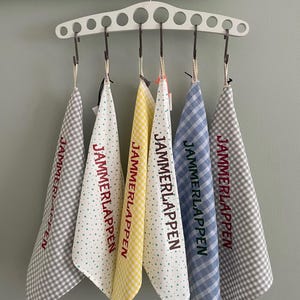 Könnte beinhalten: Ein weißer Kleiderbügel zeigt sechs gemusterte Küchentücher. Die Tücher weisen Karomuster, Tupfenmuster und Karomuster in Grau, Gelb und Blau auf. Jedes Tuch ist mit dem Text "JAMMERLAPPEN" in Rot beschriftet.