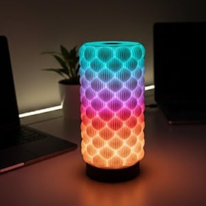 Könnte beinhalten: Zylindrische LED-Lampe mit einem strukturierten, schuppenartigen Muster. Die Lampe strahlt einen Farbverlauf aus, der von Orange an der Basis bis Blau an der Spitze reicht. Sie steht auf einem dunklen Sockel, mit einem Laptop und einer Pflanze im Hintergrund.