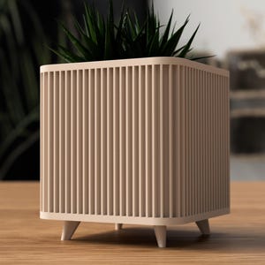 Op de afbeelding: Een vierkante, beige plantenbak met verticale lamellen. De plantenbak staat op korte, witte poten en bevat een groene plant. Het ontwerp is modern en minimalistisch, geschikt voor binnen of buiten.