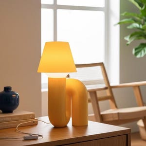 Op de afbeelding: Een gele tafellamp met een gebogen voet en een lampenkap. De lamp staat op een houten tafel naast een blauwe vaas en boeken. Het ontwerp van de lamp is minimalistisch en eigentijds.