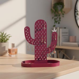 Op de afbeelding: Een bordeauxrode cactusvormige sieradenorganizer met een dienbladbasis. De organizer heeft meerdere gaten voor oorbellen en een klein kroontje. Verschillende paar oorbellen hangen aan de organizer. De organizer staat op een lichtgekleurd houten oppervlak.