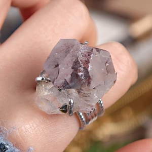 May include: A silver ring featuring a large, multifaceted crystal with a mix of clear and purple hues. The crystal has dark red inclusions. The ring's band has a twisted design.