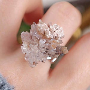 May include: A ring featuring a cluster of light brown and clear crystals. The crystals are arranged in a textured, natural formation, set on a silver band. The ring is held by a person.