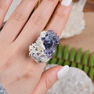 May include: A raw fluorite ring featuring a cluster of purple and white crystals. The ring showcases the natural beauty of the mineral, with a rough, textured appearance. The gemstone is set on a simple band, highlighting the unique stone.