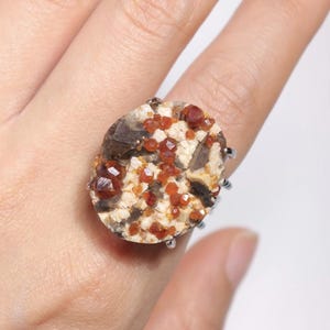 May include: A unique ring featuring a large, oval-shaped stone with a speckled beige and brown pattern. The stone is adorned with numerous small, reddish-orange crystals. The ring has a silver-colored band.