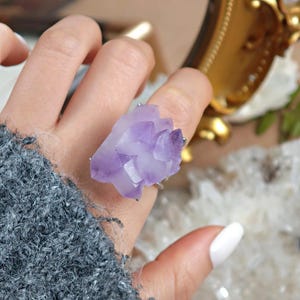 May include: A silver ring featuring a large, raw amethyst crystal. The crystal is a light purple color and has a cluster of pointed formations. The ring is set on a finger, showcasing the natural beauty of the gemstone.