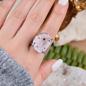 May include: A silver ring featuring a large, oval-shaped, light pink geode stone. The stone has a crystalline interior with dark brown inclusions. The ring's setting has a textured band and four prongs securing the stone.
