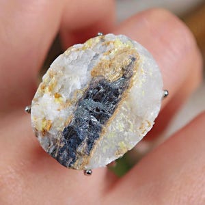 May include: A unique ring featuring a natural stone with a mix of white, black, yellow, and brown colors. The stone is mounted on a silver-colored band with four small prongs holding the stone in place. The ring is a statement piece.