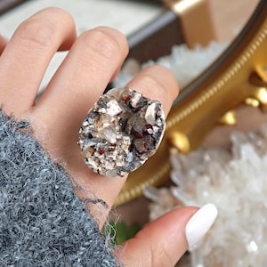 May include: A unique ring featuring a large, irregularly shaped stone with a mix of colors. The stone has a rough, textured surface with shades of gray, white, and reddish-brown. The ring is a statement piece, perfect for adding a natural, earthy element to any outfit.