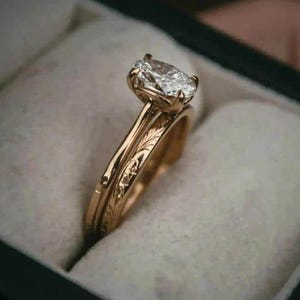 May include: A gold engagement ring featuring a large, clear, oval-cut diamond. The ring has a delicate band with engraved floral details. The ring is displayed in a dark green box with a white interior.