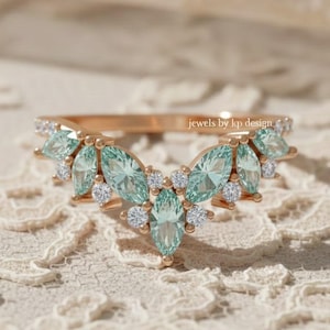 Puede incluir: Un anillo de oro rosa con un diseño curvo, con piedras preciosas azul claro en forma de marquesa y pequeñas piedras blancas. El anillo está sobre una superficie de encaje, con el texto "jewels by kp design".