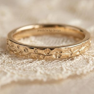 May include: A gold-colored ring with a floral and leaf pattern. The ring has a band with detailed carvings of flowers and foliage. The text "jewels by kp design" is visible on the ring. The ring is set against a lace background.