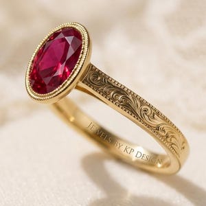 May include: A gold ring with an oval, deep red gemstone. The ring's band is engraved with a floral pattern, and the text "JEWELS BY KP DESIGN" is visible. The ring is displayed on a soft, off-white surface.