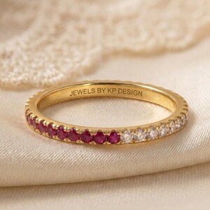 Peut inclure: Une bague en or avec une rangée de petits rubis rouges ronds et de diamants blancs transparents sertis sur le bord extérieur. L'intérieur de l'anneau porte l'inscription JEWELS BY KP DESIGN gravée dans une police sans empattement.