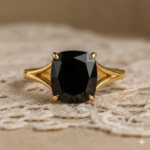 Op de afbeelding: Een gouden ring met een grote, vierkant geslepen zwarte edelsteen. De ring heeft een gespleten schachtontwerp en vier klauwen die de steen vasthouden. De ring staat tegen een neutrale, getextureerde achtergrond.